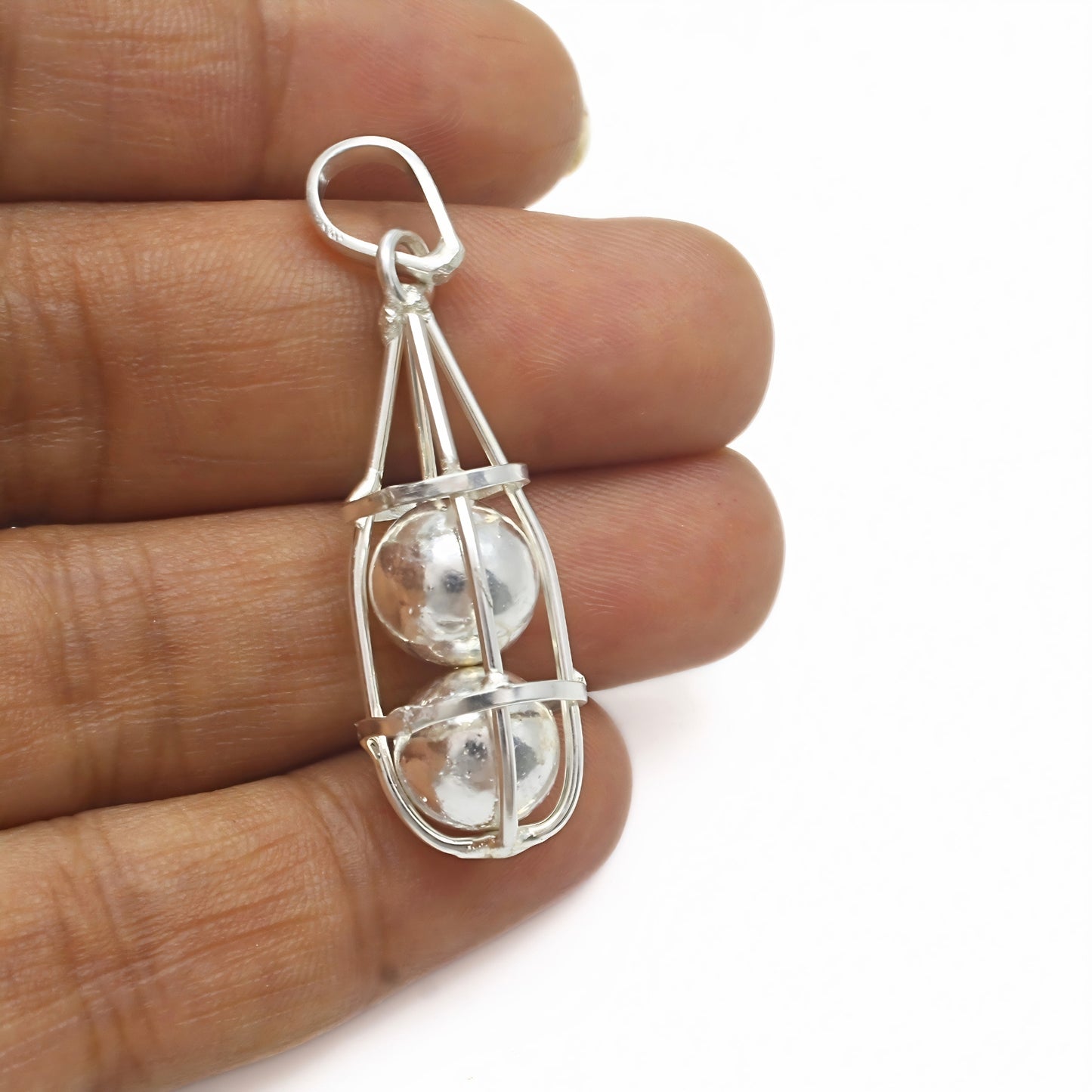 A silver cage pendant held between fingers, enclosing two stacked silver spheres against a plain white background.