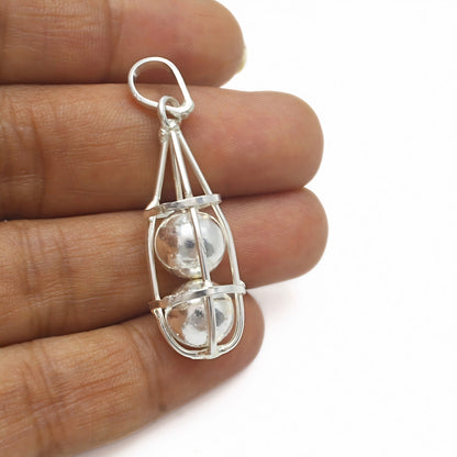 A silver cage pendant held between fingers, enclosing two stacked silver spheres against a plain white background.